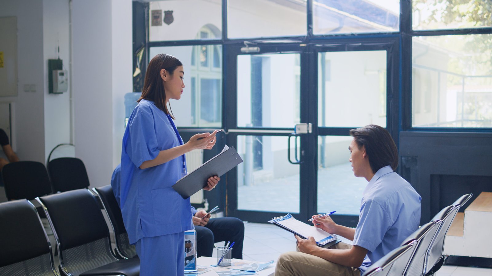 assistant-standing-hospital-counter-desk-writing-medical-expertise-clipboard-asian-patient-calls-nurse-help-him-with-insurance-information-checkup-visit-waiting-room