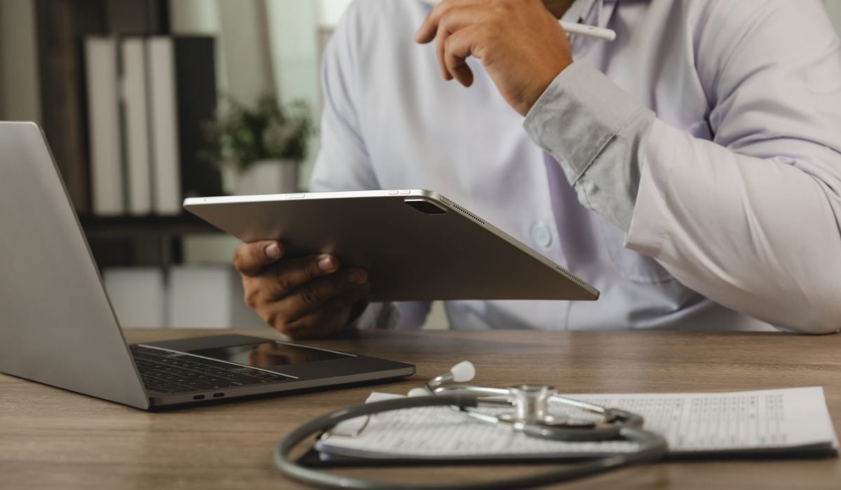 Healthcare professional reviewing patient records on a tablet beside a laptop and stethoscope at a medical desk, illustrating clinical documentation, digital healthcare workflow, patient information management, and modern medical administration.